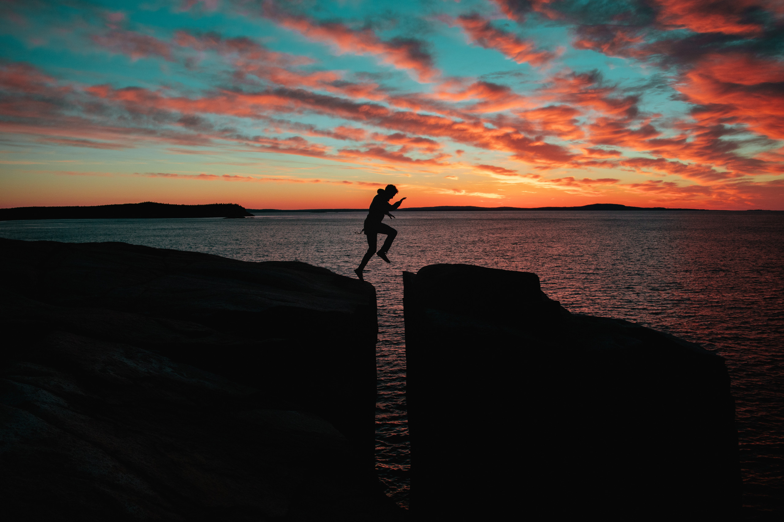 A man is jumping a gap between to rocks which are very high up. The shot is at sunset and getting dark.