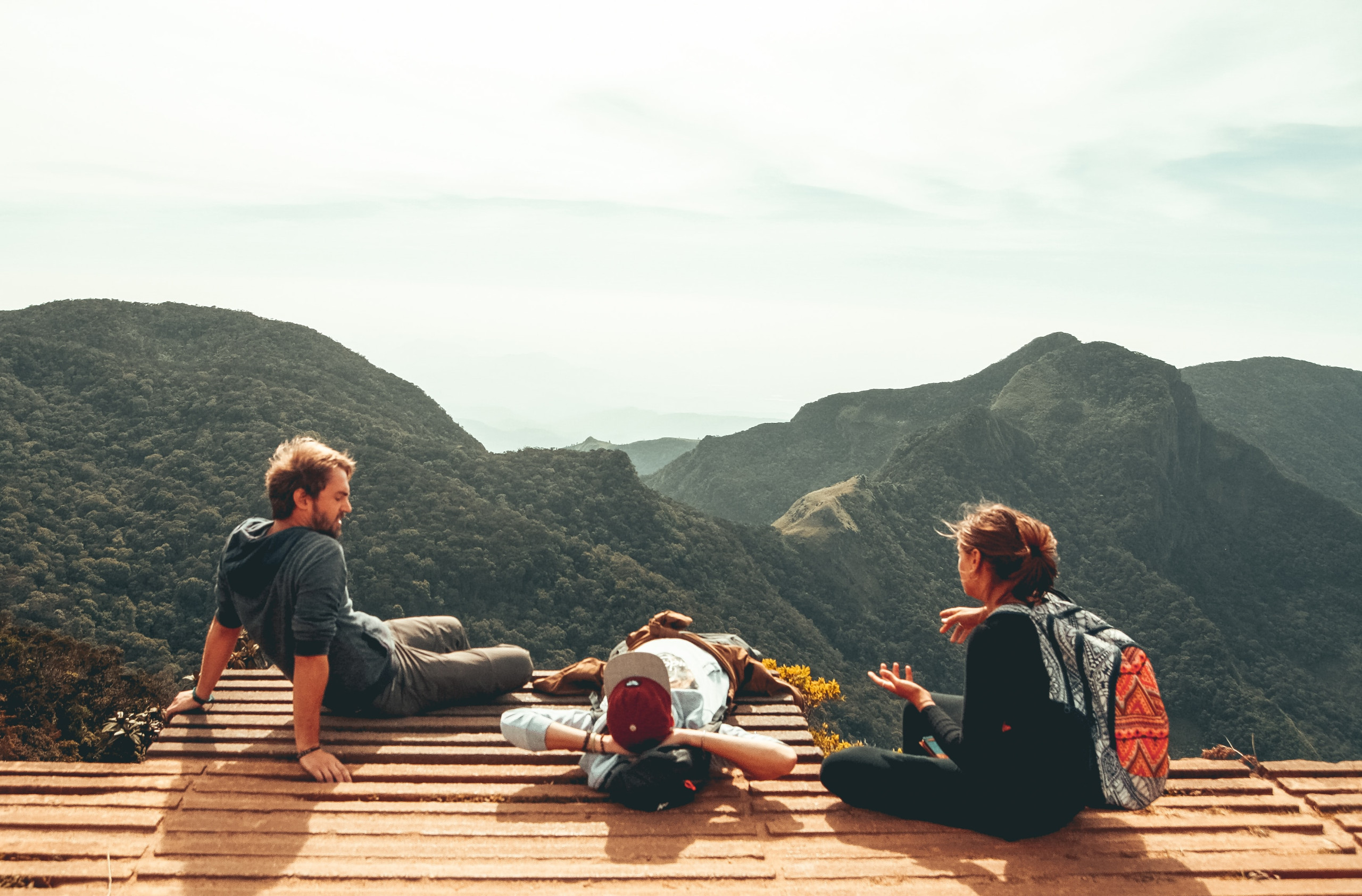 Three people sitting and the one in the middle lying down as they look over the mountain range during summer.